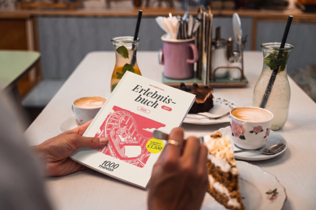 Eine Person hält das 1000things Erlebnisbuch Wien 2027 in den Händen. Im Hintergrund steht ein gedeckter Cafétisch mit Kaffee, Eistee und Kuchen.