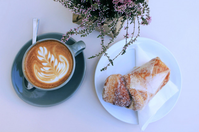 Vogelsicht auf einem creme-weißen Tisch. Links im Bild ein Kaffee mit Latte Art in grauem Geschirr, rechts auf einem weißen Teller und auf einer Serviette ein Cruffin mit Zimt und Zucker.