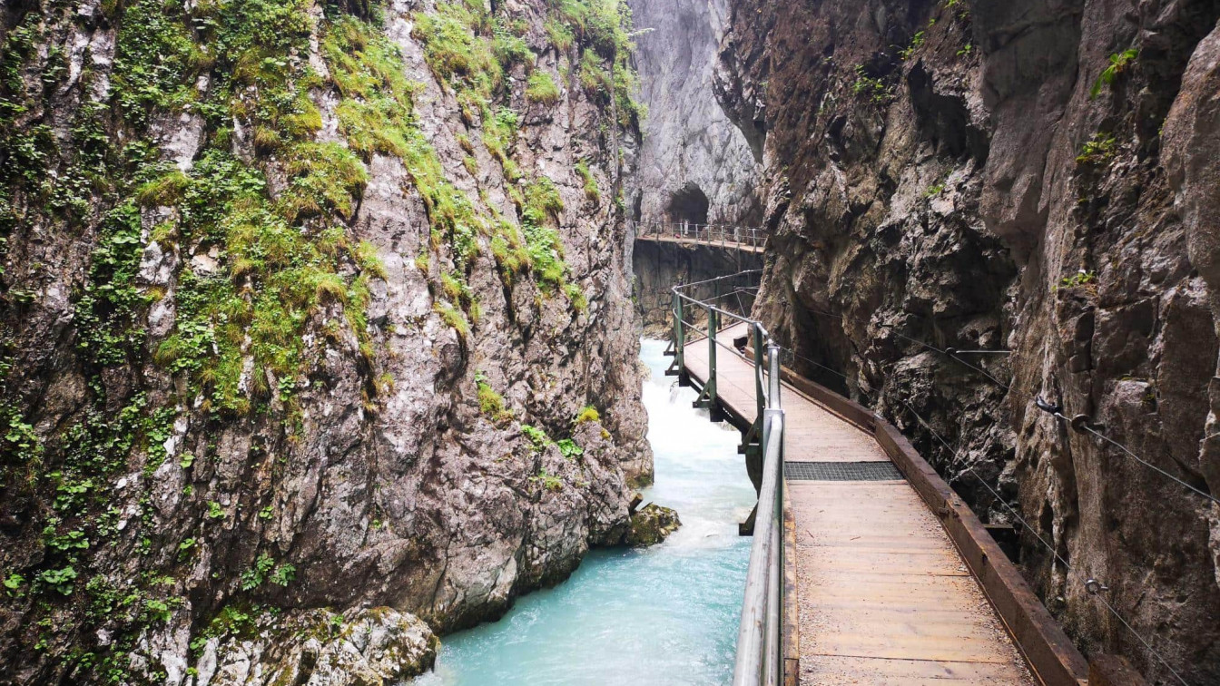 Ein schmaler Steg aus Holz und Metall führt durch die beeindruckende Leutascher Geisterklamm, flankiert von steilen Felswänden, an denen Moos und Pflanzen wachsen. Unter dem Steg fließt türkisfarbenes Wasser durch die enge Schlucht. Die Szenerie wirkt wild und geheimnisvoll, mit einem Tunnel im Hintergrund.