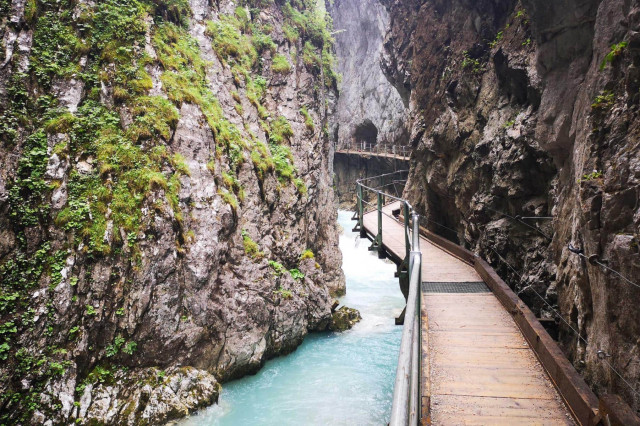 Ein schmaler Steg aus Holz und Metall führt durch die beeindruckende Leutascher Geisterklamm, flankiert von steilen Felswänden, an denen Moos und Pflanzen wachsen. Unter dem Steg fließt türkisfarbenes Wasser durch die enge Schlucht. Die Szenerie wirkt wild und geheimnisvoll, mit einem Tunnel im Hintergrund.